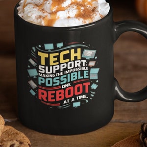 Puede incluir: Taza de café negra con crema batida blanca y un chorrito de caramelo. La taza tiene un gráfico que dice "Tech Support: Making the impossible possible one reboot at a time."
