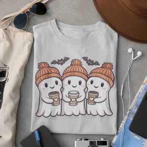 K&ouml;nnte beinhalten: Ein graues T-Shirt mit einem Motiv von drei Cartoon-Geistern, die orangefarbene Strickm&uuml;tzen tragen und Tassen Kaffee halten. Die Geister l&auml;cheln und haben Herzen auf ihren Tassen. &Uuml;ber den Geistern befinden sich zwei Flederm&auml;use.