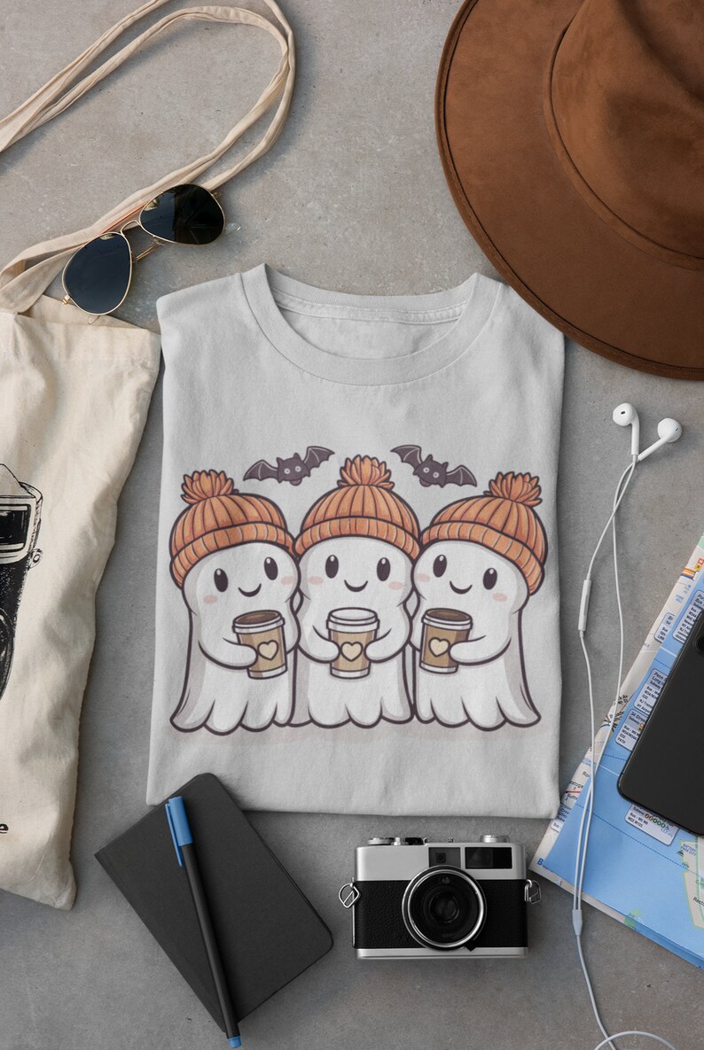 K&ouml;nnte beinhalten: Ein graues T-Shirt mit einem Motiv von drei Cartoon-Geistern, die orangefarbene Strickm&uuml;tzen tragen und Tassen Kaffee halten. Die Geister l&auml;cheln und haben Herzen auf ihren Tassen. &Uuml;ber den Geistern befinden sich zwei Flederm&auml;use.