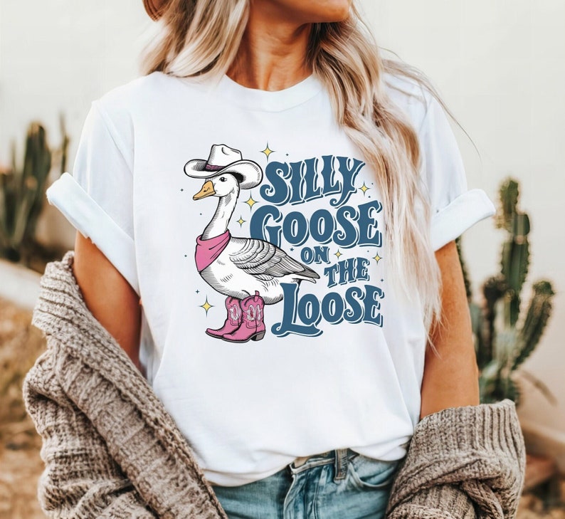 May include: White t-shirt with a cartoon goose wearing a cowboy hat and boots. The text "Silly Goose on the Loose" is printed on the shirt in blue.