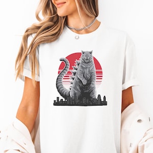 May include: White t-shirt with a graphic of a gray cat with a red sun and city skyline in the background.