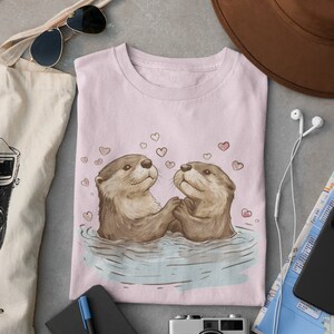 May include: A light pink t-shirt featuring a charming illustration of two otters holding hands in water, surrounded by hearts. The otters are depicted in shades of brown and tan. The shirt is folded and surrounded by travel accessories.