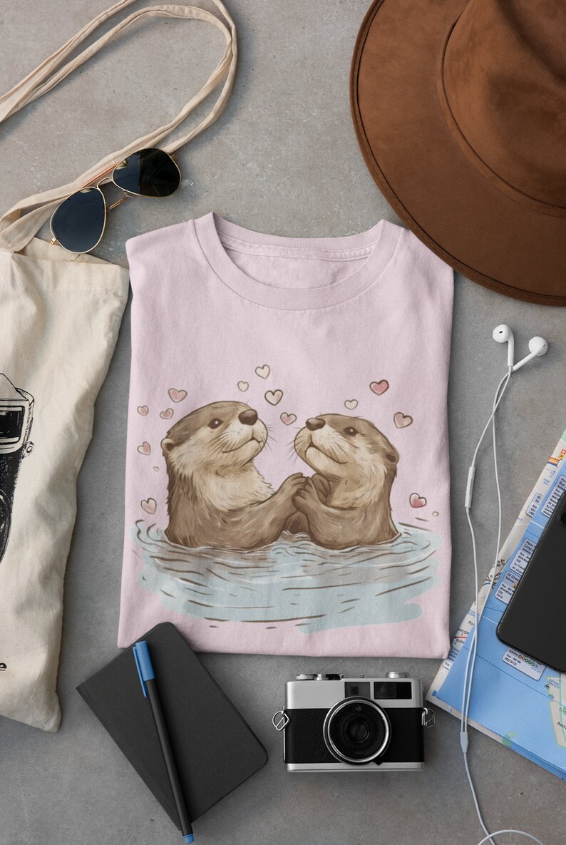 May include: A light pink t-shirt featuring a charming illustration of two otters holding hands in water, surrounded by hearts. The otters are depicted in shades of brown and tan. The shirt is folded and surrounded by travel accessories.
