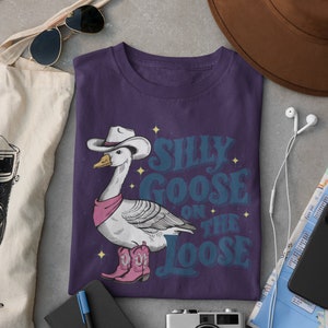 May include: Purple t-shirt with a cartoon goose wearing a cowboy hat and boots. The text "Silly Goose on the Loose" is printed on the shirt.