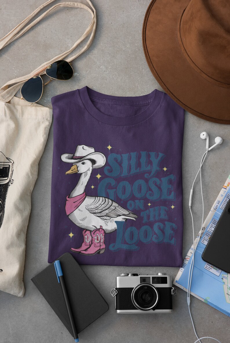 May include: Purple t-shirt with a cartoon goose wearing a cowboy hat and boots. The text "Silly Goose on the Loose" is printed on the shirt.