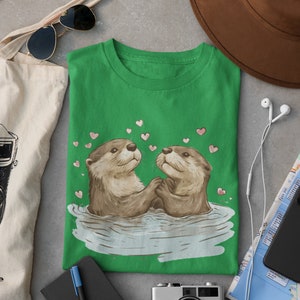 May include: Green t-shirt featuring a graphic of two otters holding hands in water, surrounded by pink hearts. The shirt is part of a collection of apparel and accessories, including a tote bag, sunglasses, and a camera.