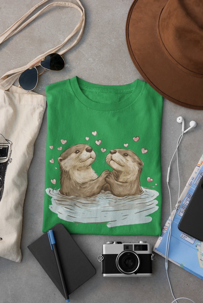 May include: Green t-shirt featuring a graphic of two otters holding hands in water, surrounded by pink hearts. The shirt is part of a collection of apparel and accessories, including a tote bag, sunglasses, and a camera.