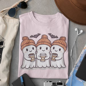 K&ouml;nnte beinhalten: Rosafarbenes T-Shirt mit einem Motiv von drei Cartoon-Geistern, die orangefarbene Strickm&uuml;tzen tragen und Tassen Kaffee halten. Die Geister l&auml;cheln und haben Flederm&auml;use, die &uuml;ber ihnen fliegen.