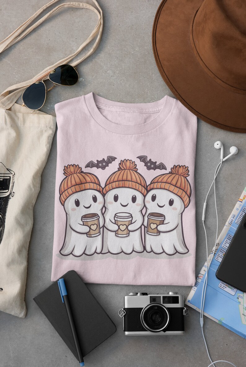 K&ouml;nnte beinhalten: Rosafarbenes T-Shirt mit einem Motiv von drei Cartoon-Geistern, die orangefarbene Strickm&uuml;tzen tragen und Tassen Kaffee halten. Die Geister l&auml;cheln und haben Flederm&auml;use, die &uuml;ber ihnen fliegen.