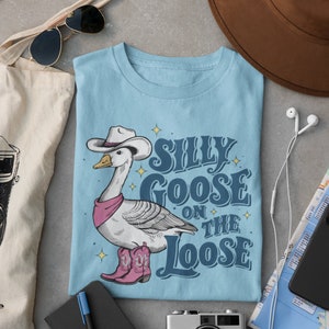 May include: A light blue t-shirt with a cartoon goose wearing a cowboy hat and boots. The text "Silly Goose on the Loose" is printed on the shirt.
