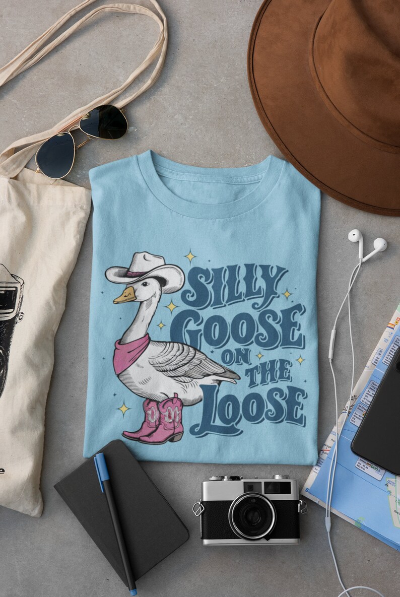 May include: A light blue t-shirt with a cartoon goose wearing a cowboy hat and boots. The text "Silly Goose on the Loose" is printed on the shirt.