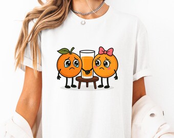 Könnte beinhalten: Weißes T-Shirt mit einem Aufdruck von zwei Orangenscheiben mit Gesichtern, eine mit einer rosa Schleife, die sich vor einem Glas Orangensaft die Hände halten. Die Orangenscheiben weinen.