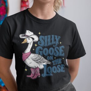 May include: Black t-shirt with a white goose wearing a cowboy hat and pink bandana. The goose is wearing pink cowboy boots. The text "Silly Goose On The Loose" is printed on the shirt in blue.