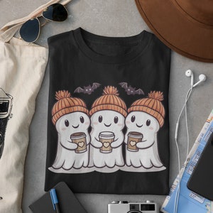 K&ouml;nnte beinhalten: Schwarzes T-Shirt mit einem Motiv von drei Cartoon-Geistern, die orangefarbene Strickm&uuml;tzen tragen und Tassen Kaffee halten. Die Geister l&auml;cheln und haben Flederm&auml;use, die &uuml;ber ihnen fliegen.