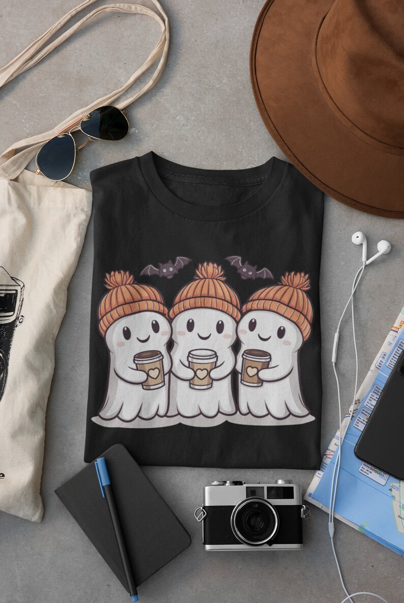 K&ouml;nnte beinhalten: Schwarzes T-Shirt mit einem Motiv von drei Cartoon-Geistern, die orangefarbene Strickm&uuml;tzen tragen und Tassen Kaffee halten. Die Geister l&auml;cheln und haben Flederm&auml;use, die &uuml;ber ihnen fliegen.