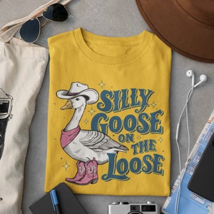 May include: Yellow t-shirt with a cartoon illustration of a goose wearing a cowboy hat and boots. The text "Silly Goose on the Loose" is printed on the shirt.