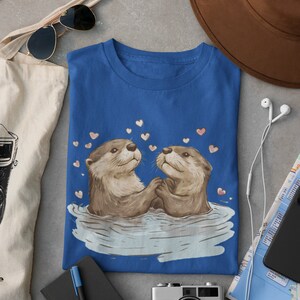 May include: Blue t-shirt featuring an illustration of two otters holding hands in water, surrounded by pink hearts. The shirt is folded on a gray surface, with a brown hat, sunglasses, and a camera nearby.