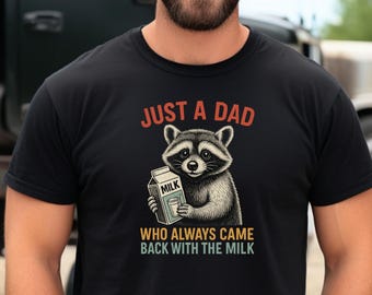 Pode incluir: Camiseta preta com a frase "JUST A DAD" em laranja acima de uma ilustração de um guaxinim segurando uma caixa de leite. Abaixo, o texto diz "WHO ALWAYS CAME BACK WITH THE MILK" em uma fonte vintage.
