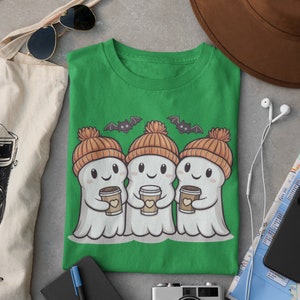 K&ouml;nnte beinhalten: Gr&uuml;nes T-Shirt mit drei Cartoon-Geistern, die orangefarbene Strickm&uuml;tzen tragen und Tassen Kaffee halten. Die Geister l&auml;cheln und haben Flederm&auml;use, die &uuml;ber ihnen fliegen.