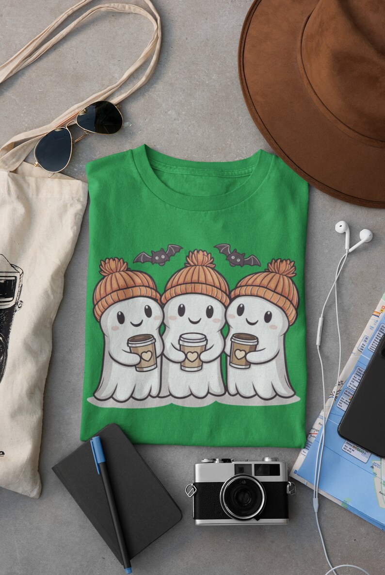 K&ouml;nnte beinhalten: Gr&uuml;nes T-Shirt mit drei Cartoon-Geistern, die orangefarbene Strickm&uuml;tzen tragen und Tassen Kaffee halten. Die Geister l&auml;cheln und haben Flederm&auml;use, die &uuml;ber ihnen fliegen.