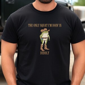 May include: Black t-shirt with a cartoon frog wearing a cowboy hat and boots. The text "The only squat I'm doin' is diddly" is printed on the shirt.