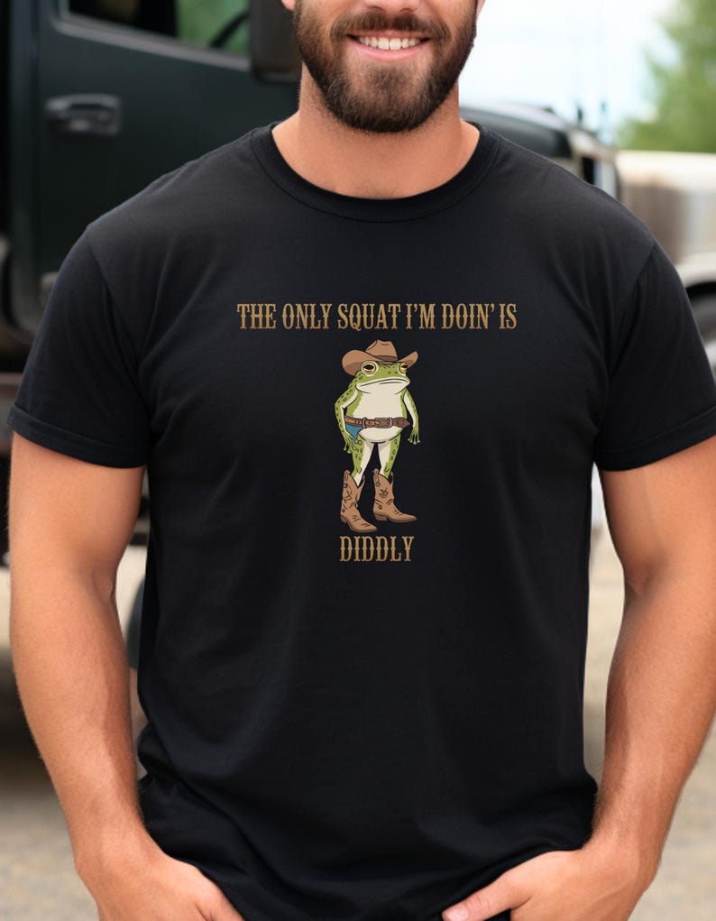 May include: Black t-shirt with a cartoon frog wearing a cowboy hat and boots. The text "The only squat I'm doin' is diddly" is printed on the shirt.