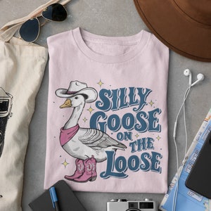 May include: A pink t-shirt with a cartoon illustration of a goose wearing a cowboy hat and boots. The text "Silly Goose on the Loose" is printed on the shirt.