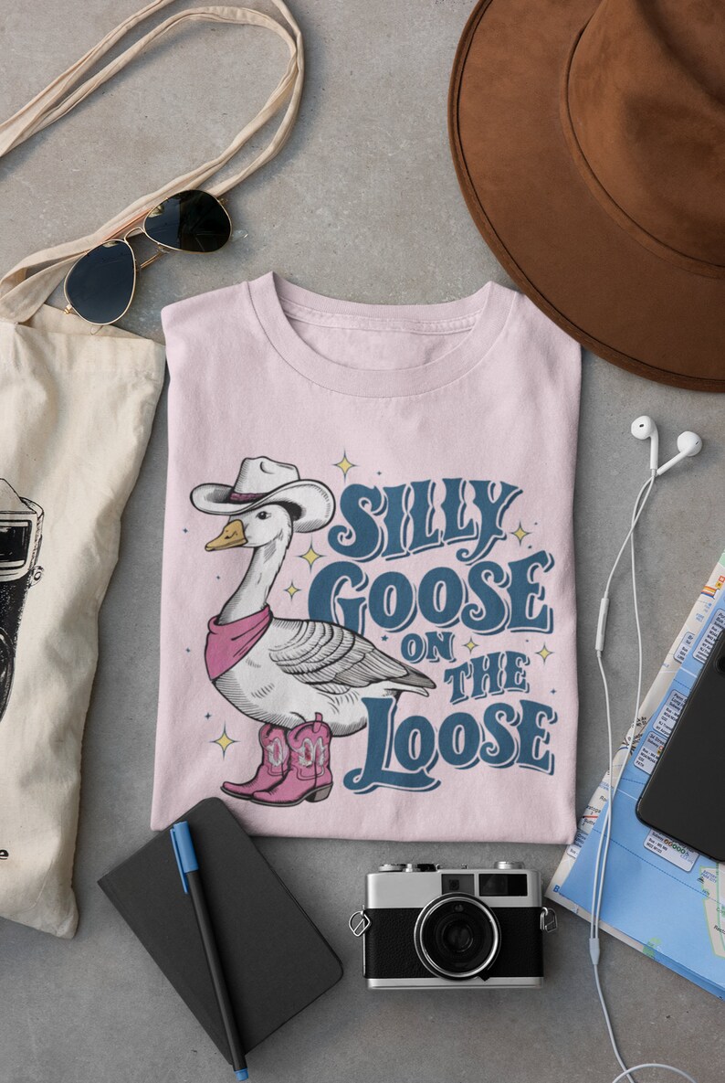 May include: A pink t-shirt with a cartoon illustration of a goose wearing a cowboy hat and boots. The text "Silly Goose on the Loose" is printed on the shirt.