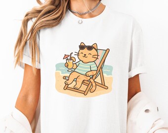 May include: White t-shirt with a cartoon illustration of a cat wearing sunglasses and relaxing on a beach chair with a drink. The cat is orange and white with a blue and white striped shirt.