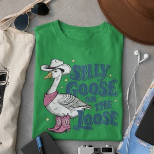 May include: Green t-shirt with a cartoon illustration of a goose wearing a cowboy hat and boots. The text "Silly Goose on the Loose" is printed on the shirt.