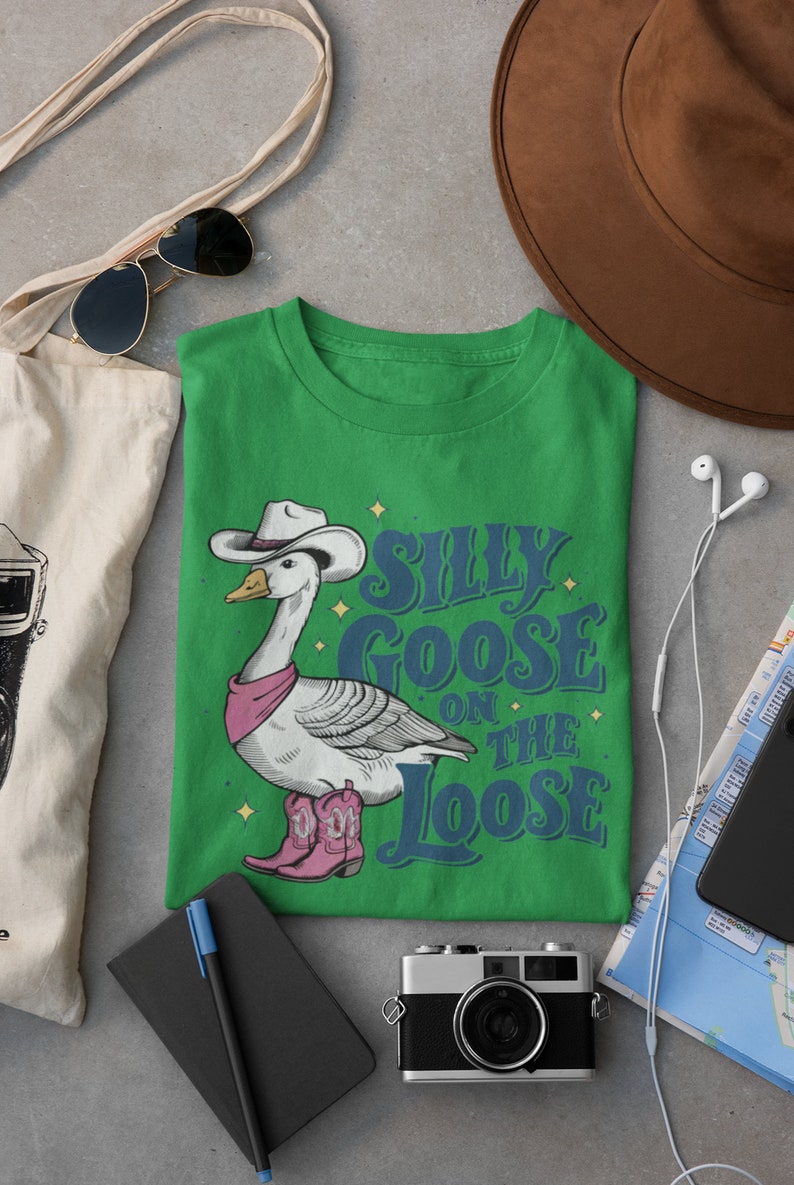 May include: Green t-shirt with a cartoon illustration of a goose wearing a cowboy hat and boots. The text "Silly Goose on the Loose" is printed on the shirt.