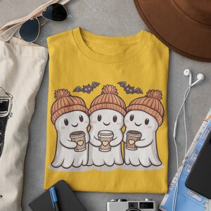K&ouml;nnte beinhalten: Ein gelbes T-Shirt mit einem Motiv von drei Geistern, die orangefarbene und braune Strickm&uuml;tzen tragen und Tassen Kaffee halten. Die Geister l&auml;cheln und &uuml;ber ihnen fliegen Flederm&auml;use.
