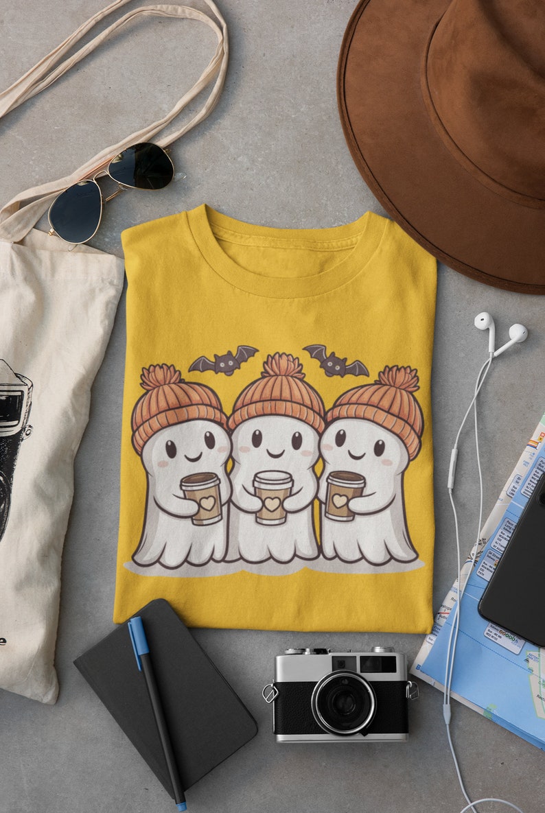 K&ouml;nnte beinhalten: Ein gelbes T-Shirt mit einem Motiv von drei Geistern, die orangefarbene und braune Strickm&uuml;tzen tragen und Tassen Kaffee halten. Die Geister l&auml;cheln und &uuml;ber ihnen fliegen Flederm&auml;use.