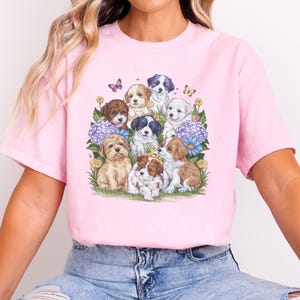 K&ouml;nnte beinhalten: Ein pinkfarbenes T-Shirt mit einem grafischen Druck einer Gruppe von Welpen, umgeben von Blumen und Schmetterlingen.