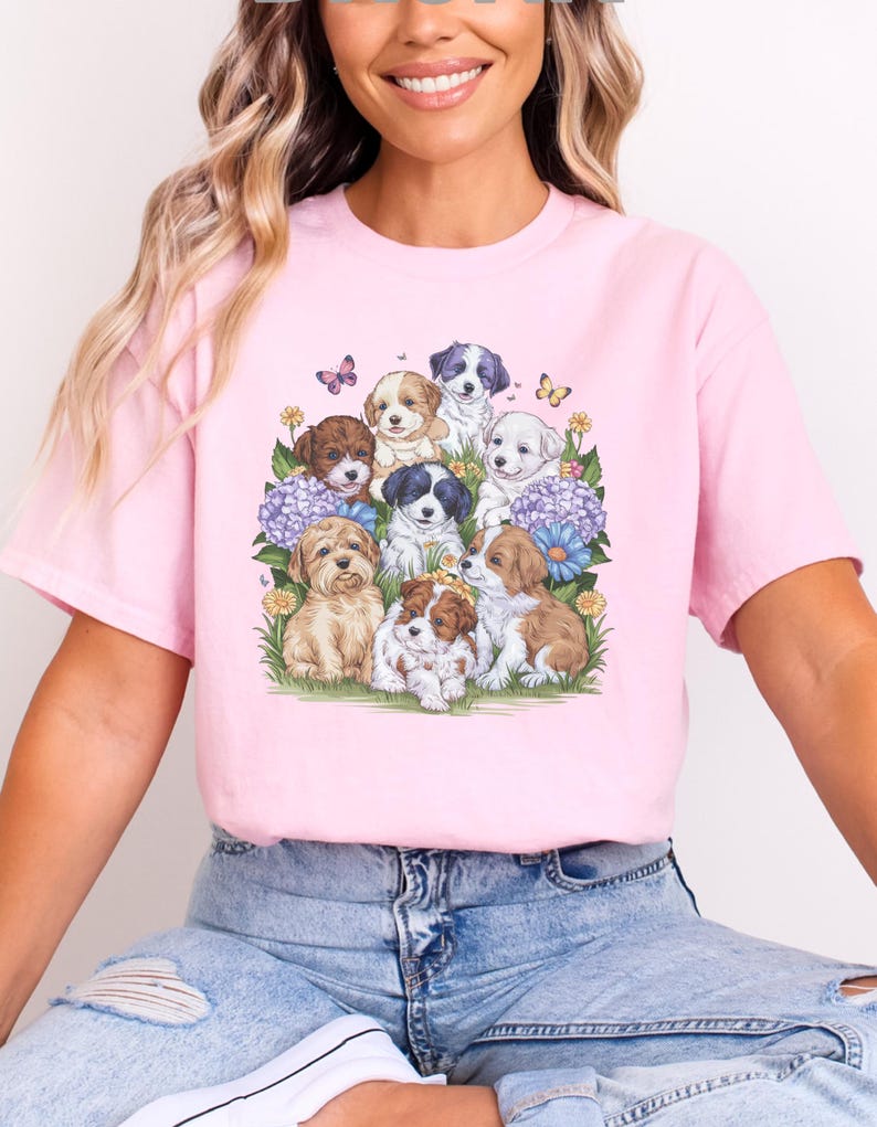 K&ouml;nnte beinhalten: Ein pinkfarbenes T-Shirt mit einem grafischen Druck einer Gruppe von Welpen, umgeben von Blumen und Schmetterlingen.