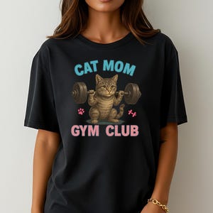 Könnte beinhalten: Schwarzes T-Shirt mit einer Grafik einer Katze, die Gewichte hebt. Der Text "CAT MOM" steht in Blau über der Katze, und "GYM CLUB" steht in Rosa darunter. Enthält rosa Pfotenabdrücke und eine Hantelgrafik.