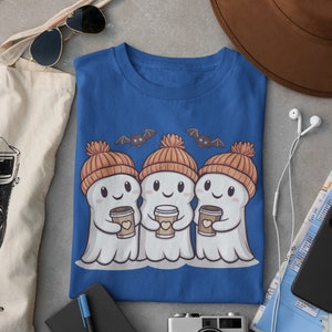 K&ouml;nnte beinhalten: Ein blaues T-Shirt mit einem Motiv von drei Geistern, die orangefarbene Strickm&uuml;tzen tragen und Tassen Kaffee halten. Die Geister l&auml;cheln und haben Flederm&auml;use, die &uuml;ber ihnen fliegen.