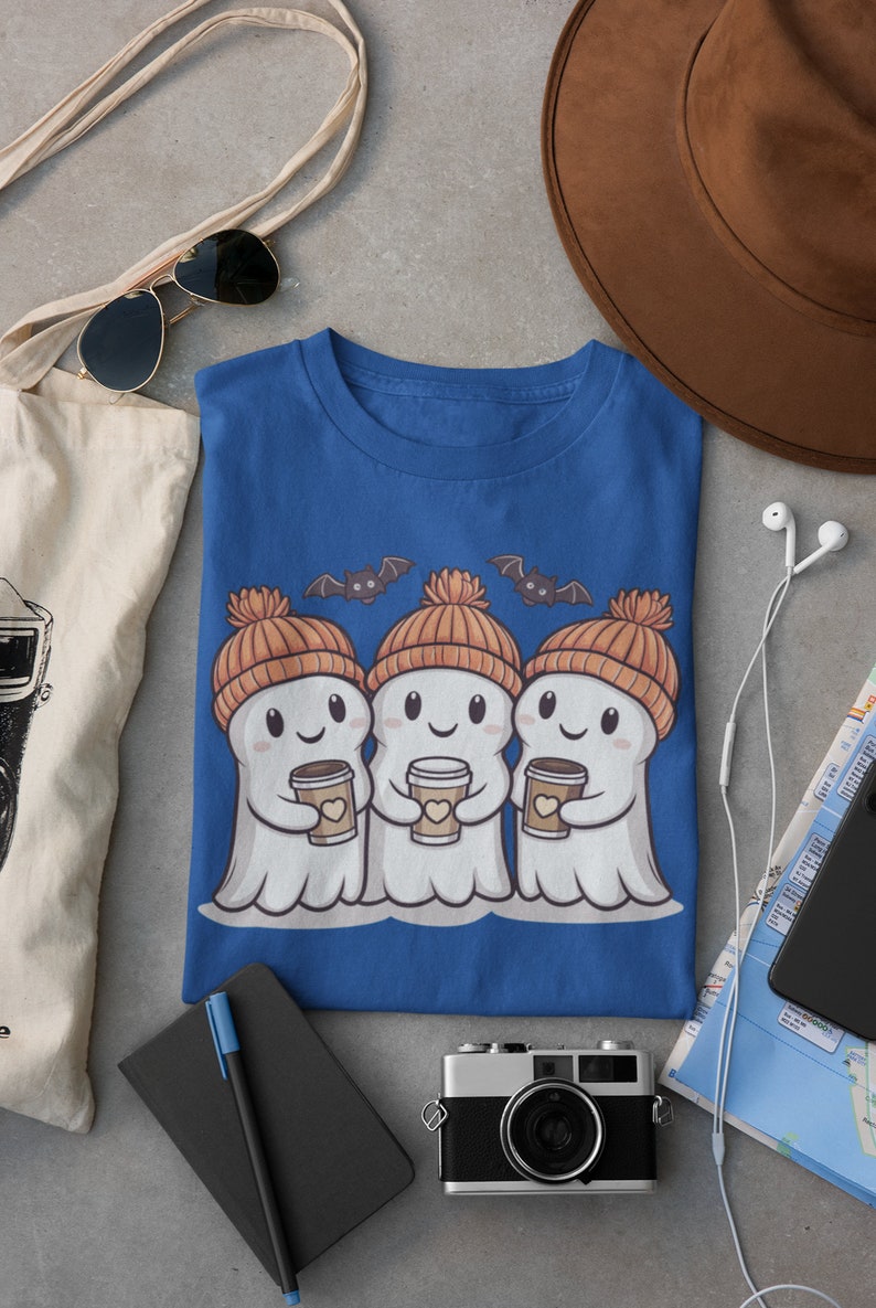K&ouml;nnte beinhalten: Ein blaues T-Shirt mit einem Motiv von drei Geistern, die orangefarbene Strickm&uuml;tzen tragen und Tassen Kaffee halten. Die Geister l&auml;cheln und haben Flederm&auml;use, die &uuml;ber ihnen fliegen.