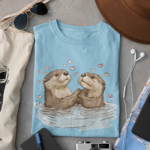May include: Light blue t-shirt featuring a charming illustration of two otters holding hands in water, surrounded by pink hearts. The otters are depicted with brown fur and expressive faces. The shirt is folded on a gray surface.