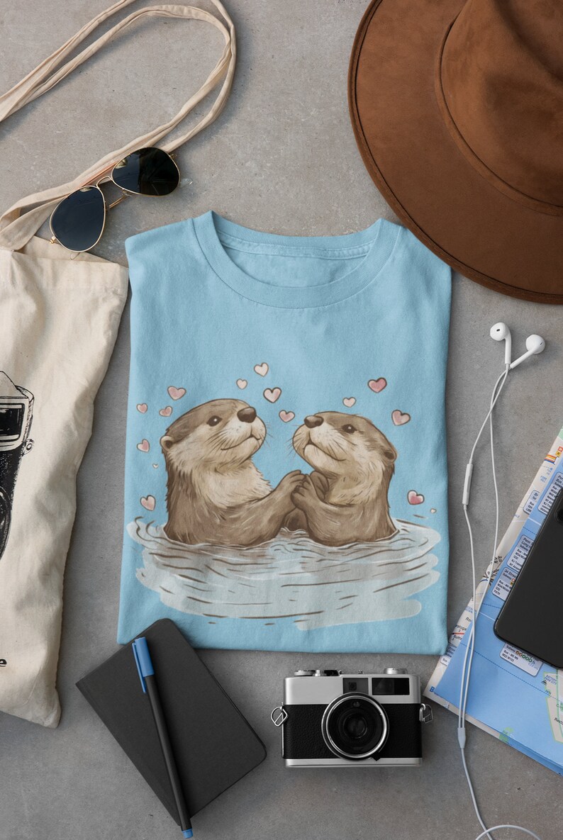 May include: Light blue t-shirt featuring a charming illustration of two otters holding hands in water, surrounded by pink hearts. The otters are depicted with brown fur and expressive faces. The shirt is folded on a gray surface.