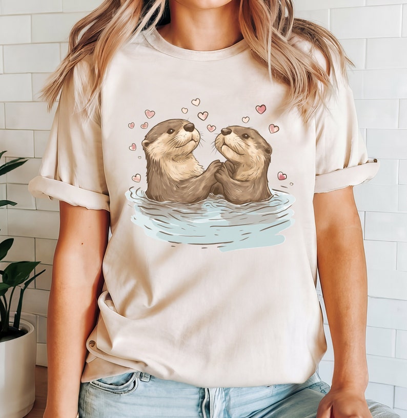 May include: Beige t-shirt featuring a charming illustration of two otters holding hands in water, surrounded by pink hearts. The otters are depicted with brown fur and are looking at each other. A cute and romantic design.