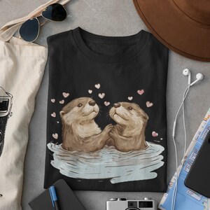 May include: Black t-shirt featuring an illustration of two otters holding hands in water, surrounded by pink hearts. The otters are brown and tan, with a light blue water effect. The shirt is displayed with accessories.