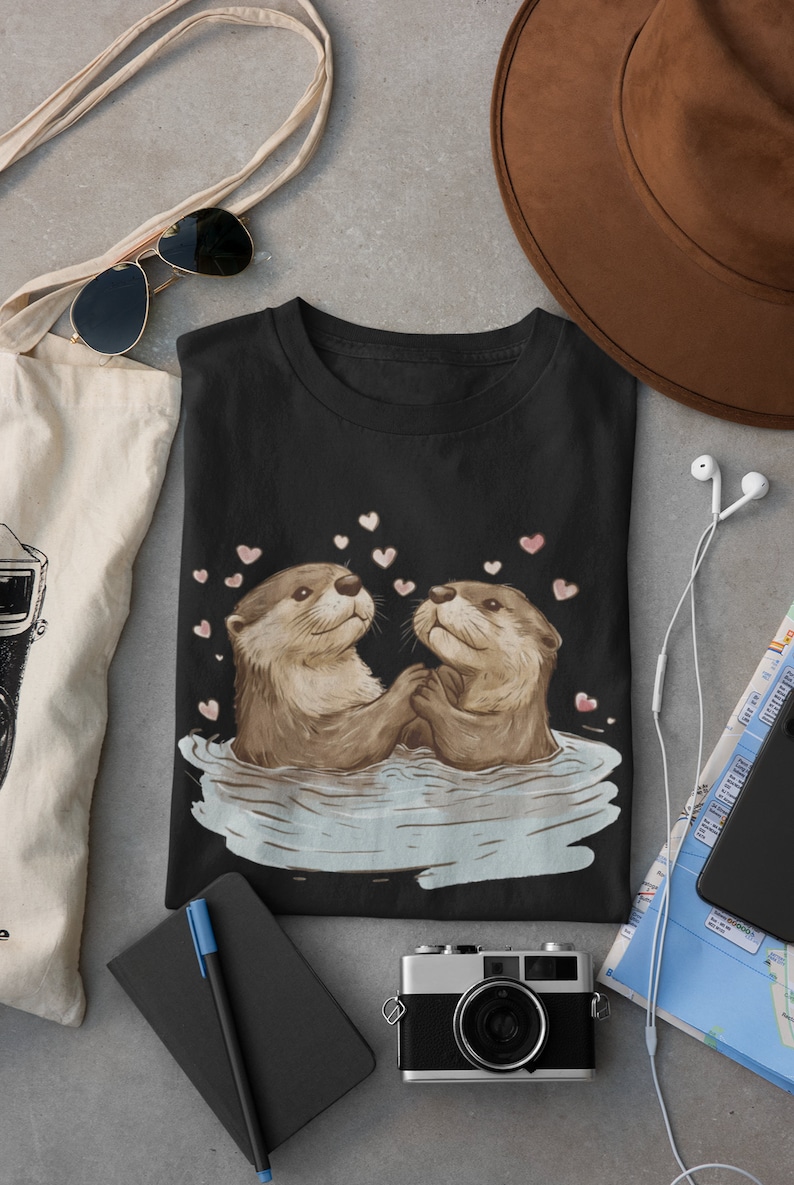 May include: Black t-shirt featuring an illustration of two otters holding hands in water, surrounded by pink hearts. The otters are brown and tan, with a light blue water effect. The shirt is displayed with accessories.