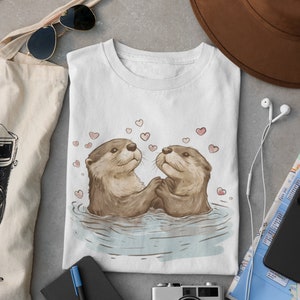 May include: White t-shirt featuring an illustration of two otters holding hands in water, surrounded by pink hearts. The otters are brown with light brown fur. The shirt is folded and placed on a gray surface, with a tote bag, sunglasses, and a camera nearby.