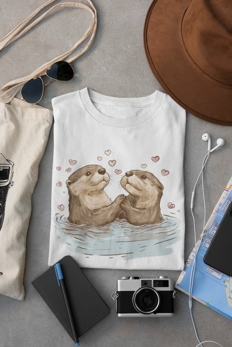 May include: White t-shirt featuring an illustration of two otters holding hands in water, surrounded by pink hearts. The otters are brown with light brown fur. The shirt is folded and placed on a gray surface, with a tote bag, sunglasses, and a camera nearby.