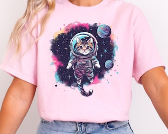 以下が含まれることがあります： 宇宙空間で宇宙服を着た猫のグラフィックがプリントされたピンクのTシャツ。猫は星と惑星に囲まれています。