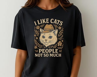 May include: Black t-shirt with a graphic of a cat wearing a hat, surrounded by floral elements. The text reads "I LIKE CATS PEOPLE NOT SO MUCH" in a serif font. The design is in shades of beige and brown.
