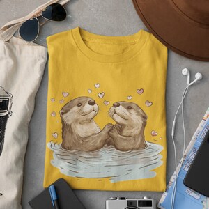 May include: Mustard yellow t-shirt featuring an illustration of two otters holding hands in water, surrounded by pink hearts. The shirt is folded on a gray surface, with a brown hat, sunglasses, and a tote bag in the background.