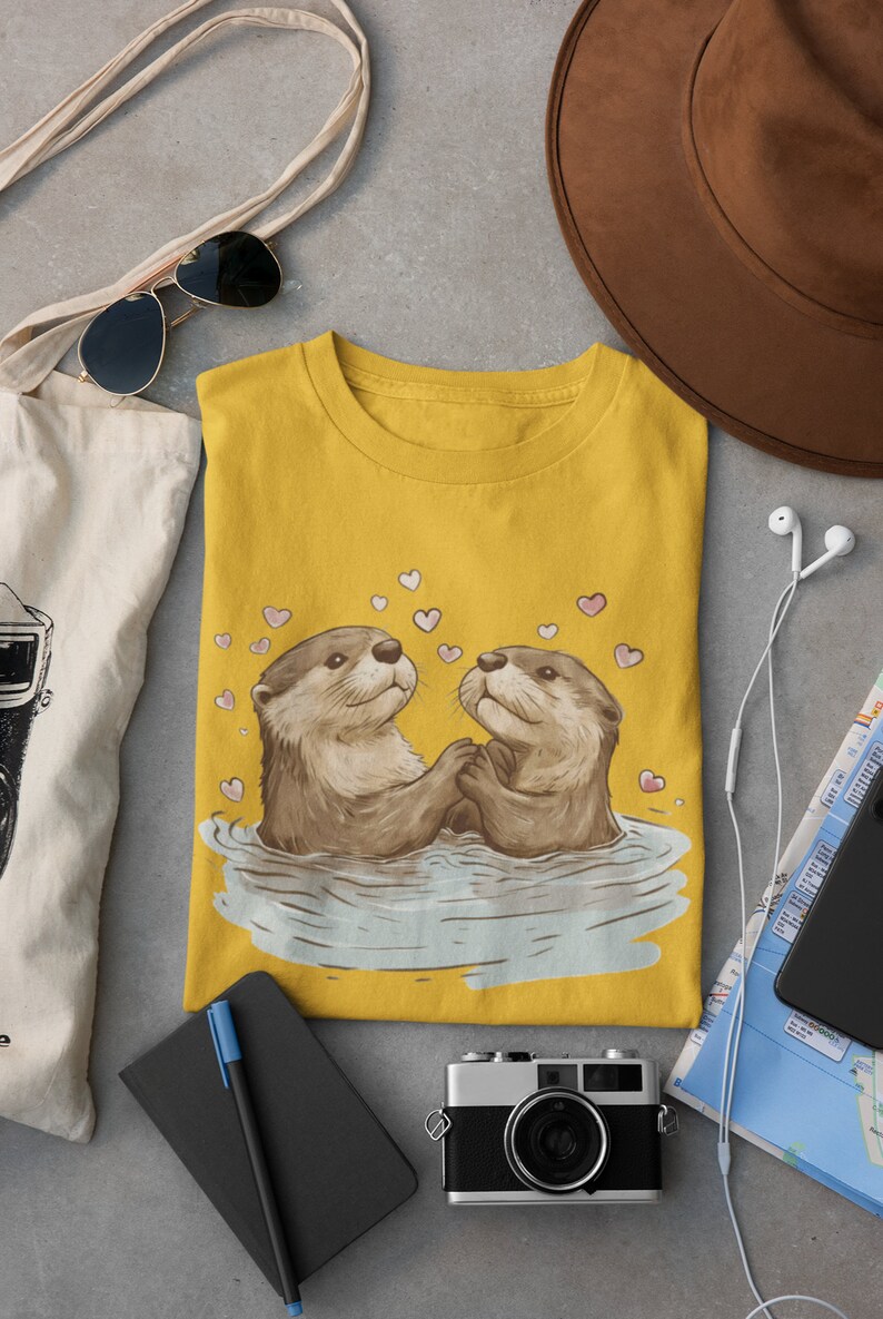 May include: Mustard yellow t-shirt featuring an illustration of two otters holding hands in water, surrounded by pink hearts. The shirt is folded on a gray surface, with a brown hat, sunglasses, and a tote bag in the background.