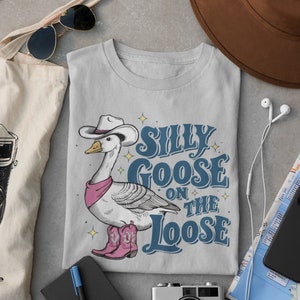 May include: A gray t-shirt with a cartoon goose wearing a cowboy hat and boots. The text "Silly Goose on the Loose" is printed on the shirt.